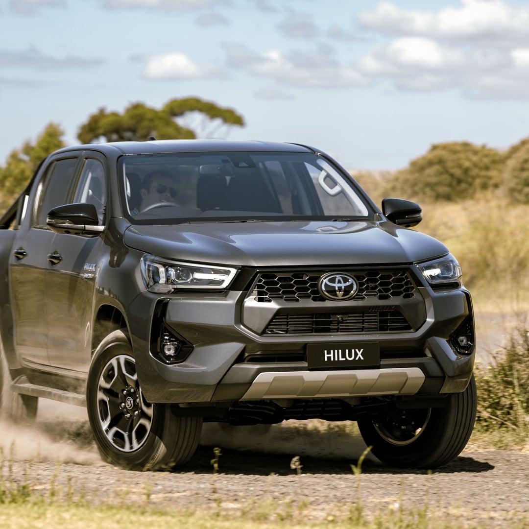 A HiLux ute with Ash Slate paint and a black, powder-coated Bull Bar drives down an unpaved road, with trees and a blue summer sky in the background.