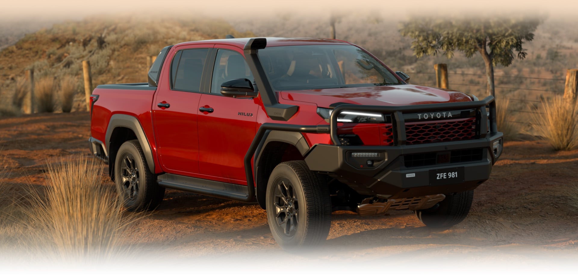 A HiLux Rogue with Feverish Red paint and a black, powder-coated Bull Bar and black Snorkel sits parked on an unpaved, red-dirt road at golden hour. In the background is a small tree, paddock fencing and Australian native grasses.