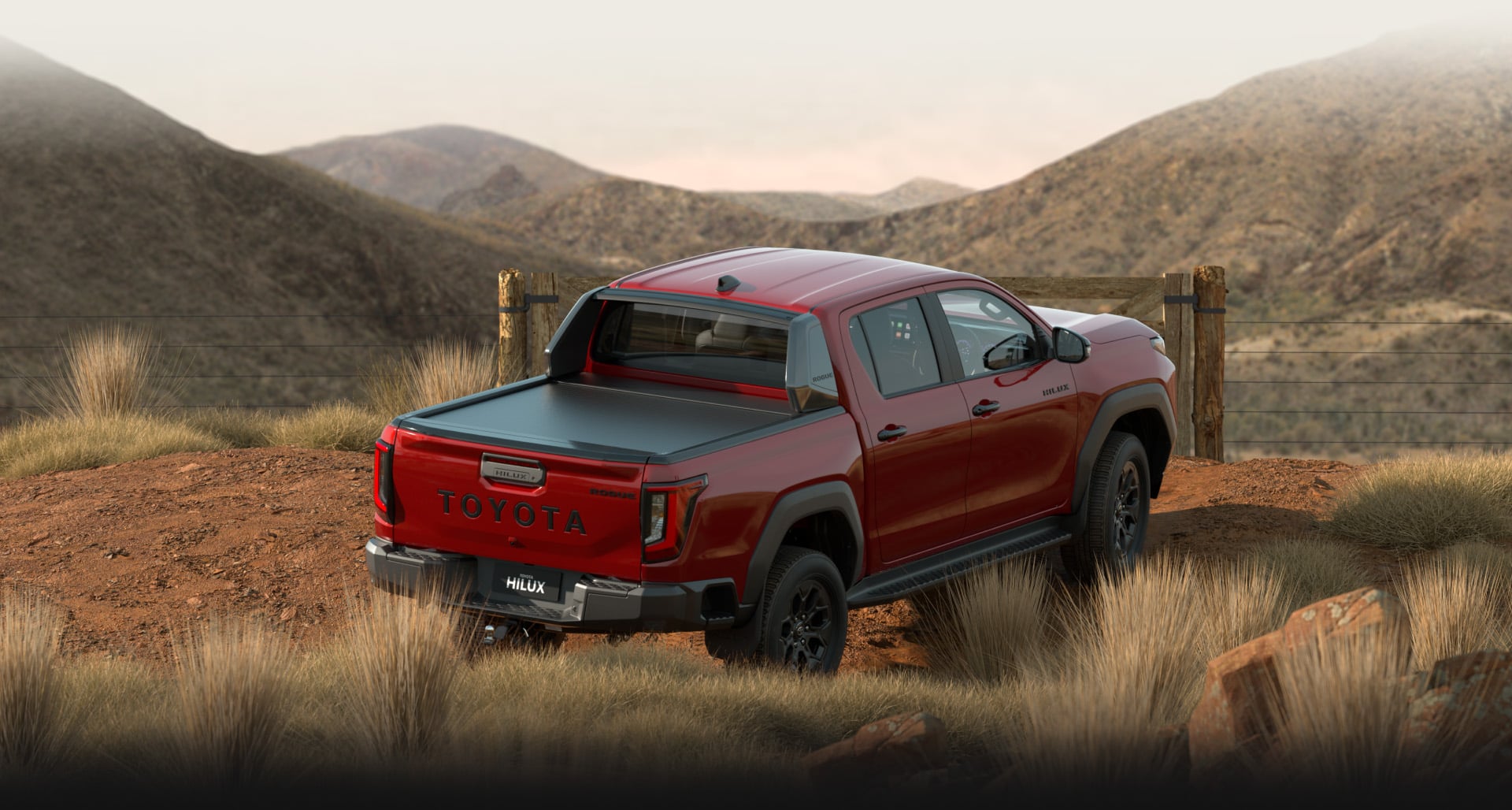 A Feverish Red HiLux Rogue is parked on a red-dirt road facing a paddock gate. The scene is set during golden hour with rolling mountains and native Australian grasses visible in the background and foreground.