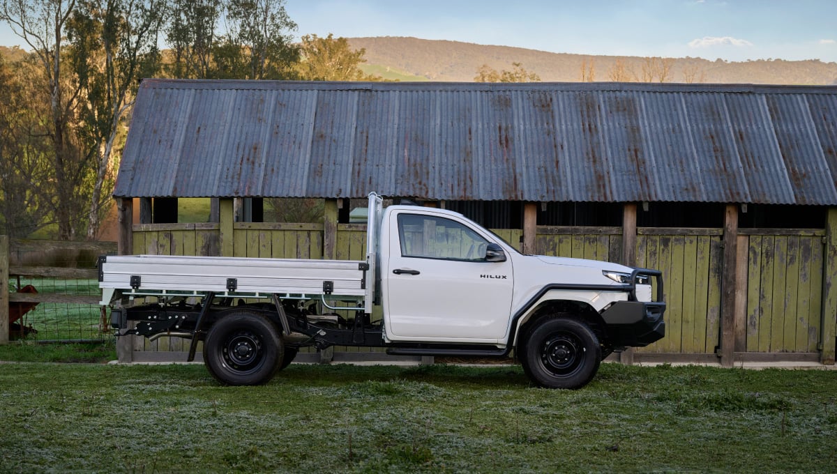 A Glacier White, single-cab HiLux WorkMate fitted with a black Bull Bar is parked in front of a rustic farm shed. The shed has a corrugated tin roof and painted, timber-panelled walls. A mountain is visible in the background.