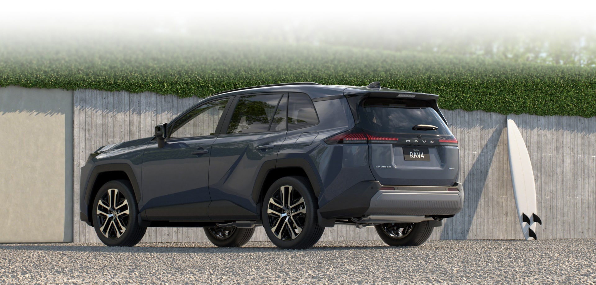 A RAV4 in Dynamic Blue is parked on a gravel surface in front of a modern concrete wall with green hedges. A white surfboard rests upright against the wall beside the vehicle, suggesting an active, coastal lifestyle.
